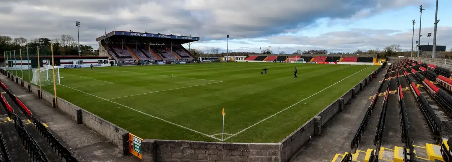 Wide-angle view of a football stadium with green grass, empty tiered stands, and a covered main stand under a cloudy sky. A few people are on the pitch near the goal area.