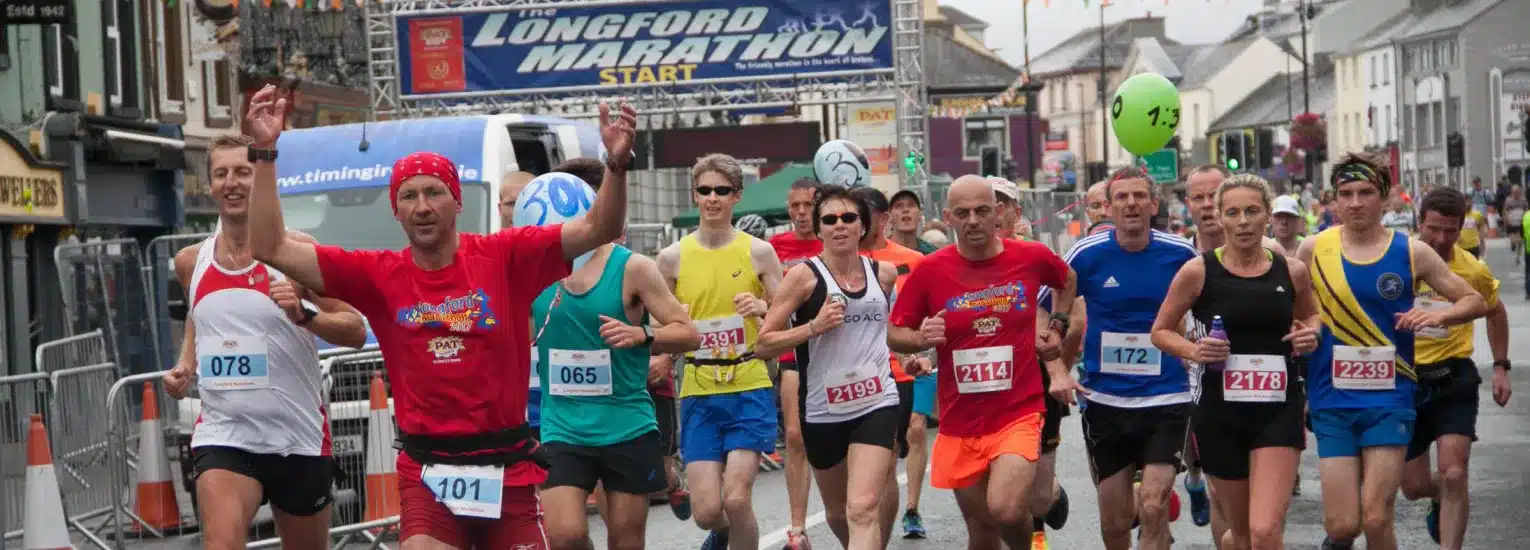 A group of runners of various ages and genders begins a race on a city street under a banner reading