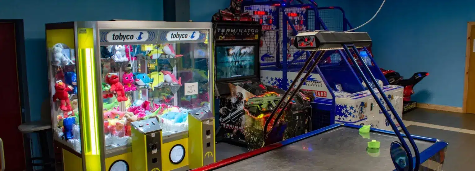 A small amusement arcade room featuring a grab machine filled with colourful soft toys, an air hockey table in the foreground, and basketball and racing arcade games in the background.
