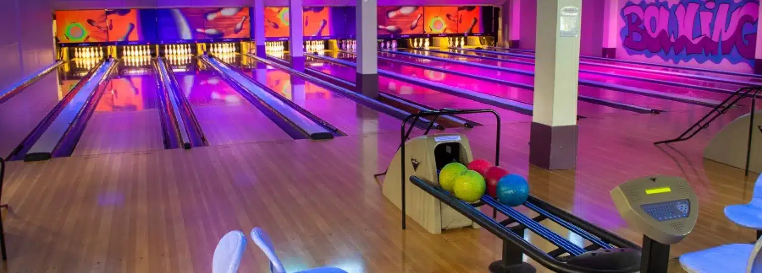 Colourful bowling alley with multiple lanes, neon and purple lighting, a rack of colourful bowling balls, scoring screens, and chairs. Walls are decorated with vibrant artwork and the word "Bowling" is painted on the right.