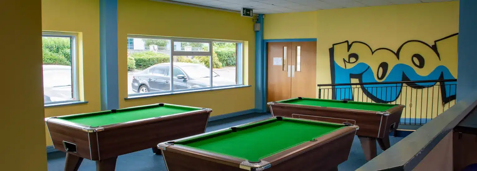 A brightly lit room with three pool tables, large windows showing parked cars outside, yellow and blue walls, and a wall mural featuring abstract shapes and the word "pool".