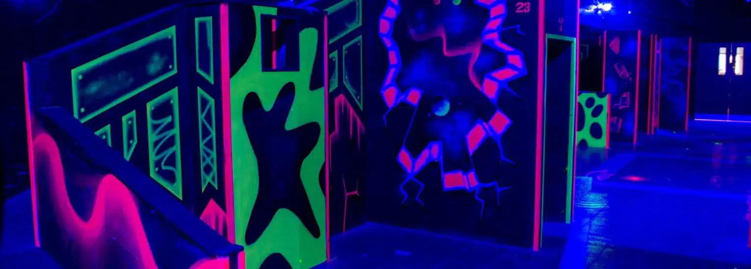 A neon-lit laser tag arena featuring black walls with bright green and pink designs, glowing obstacles, and blue lighting throughout the space.