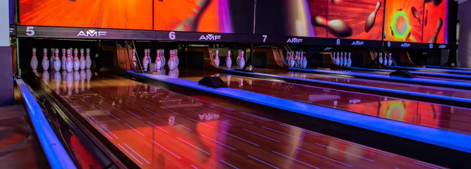 Four brightly lit bowling lanes with pins set up at the end, colourful reflections on the polished wooden floor, and vibrant images displayed on screens above each lane.