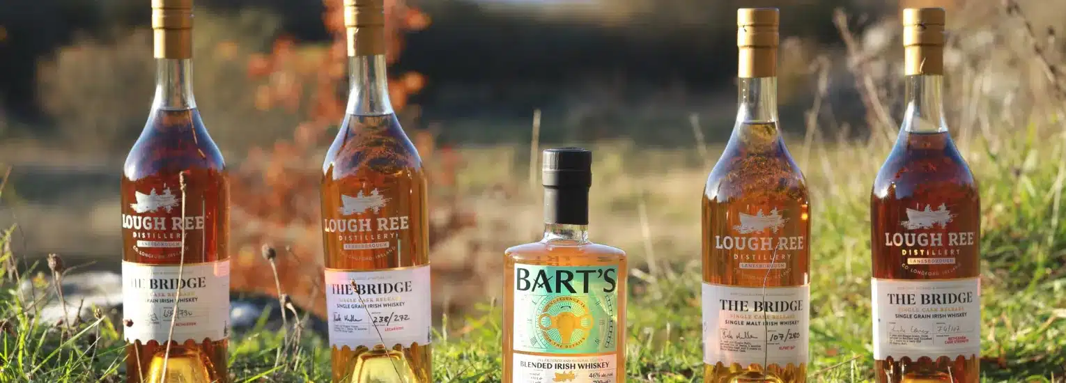 Five bottles of whisky, four labelled "Lough Ree The Bridge" and one in the centre labelled "Bart's," are displayed outdoors in sunlight on grass, with a blurred natural background.