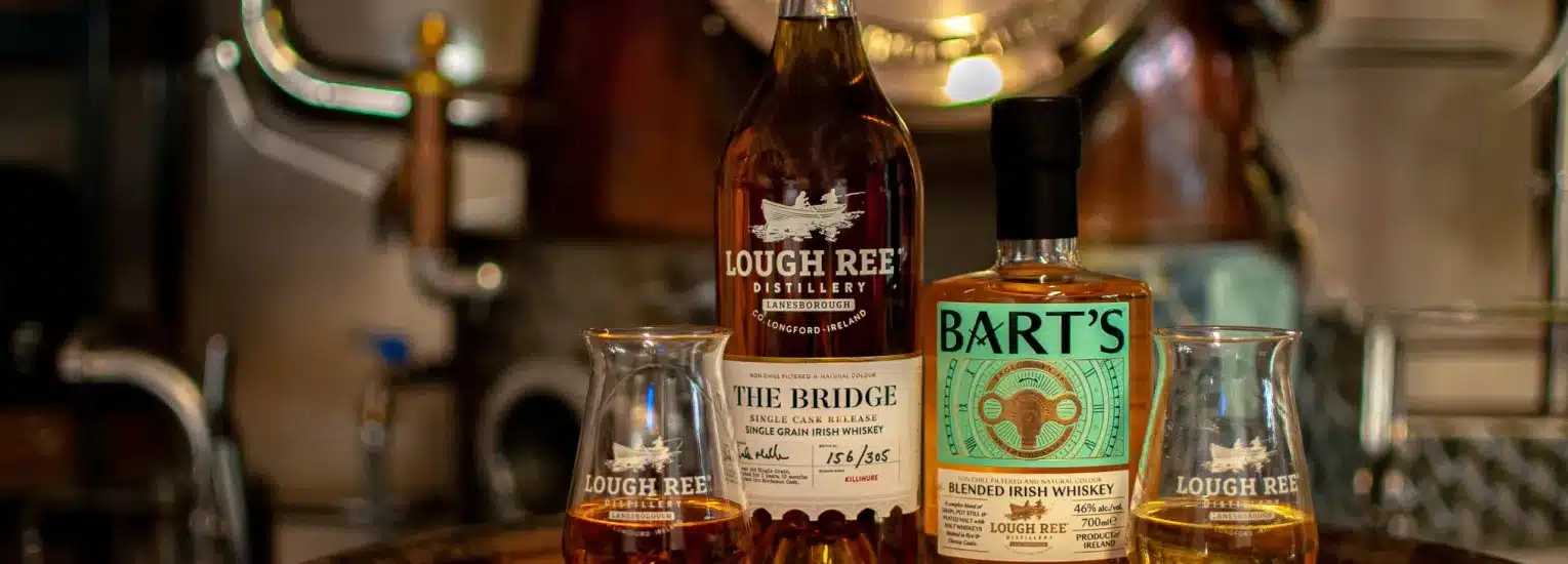 Two bottles of Irish whisky, one labelled "Lough Ree The Bridge" and the other "Bart’s," stand on a bar next to two whisky glasses filled with amber liquid, with distillery equipment blurred in the background.