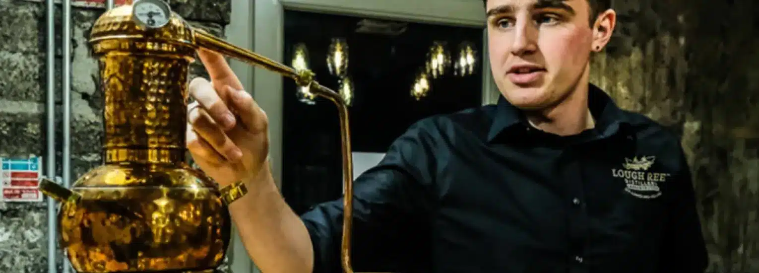 A man in a black shirt gestures towards a shiny copper still in a distillery, with stone walls and hanging lights visible in the background.