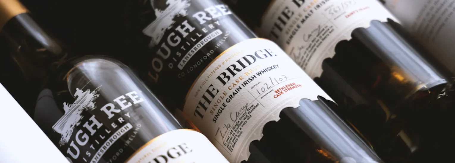 Close-up of several Lough Ree Distillery whisky bottles, with labels reading "The Bridge Single Grain Irish Whisky" lying side by side in a box, showing handwritten batch details on the labels.