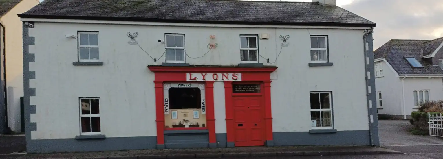 A white building with grey trim features a red shopfront labelled “LYONS.” The large front window displays various items, and the door is bright red. The building has two chimneys and four windows on the upper floor.