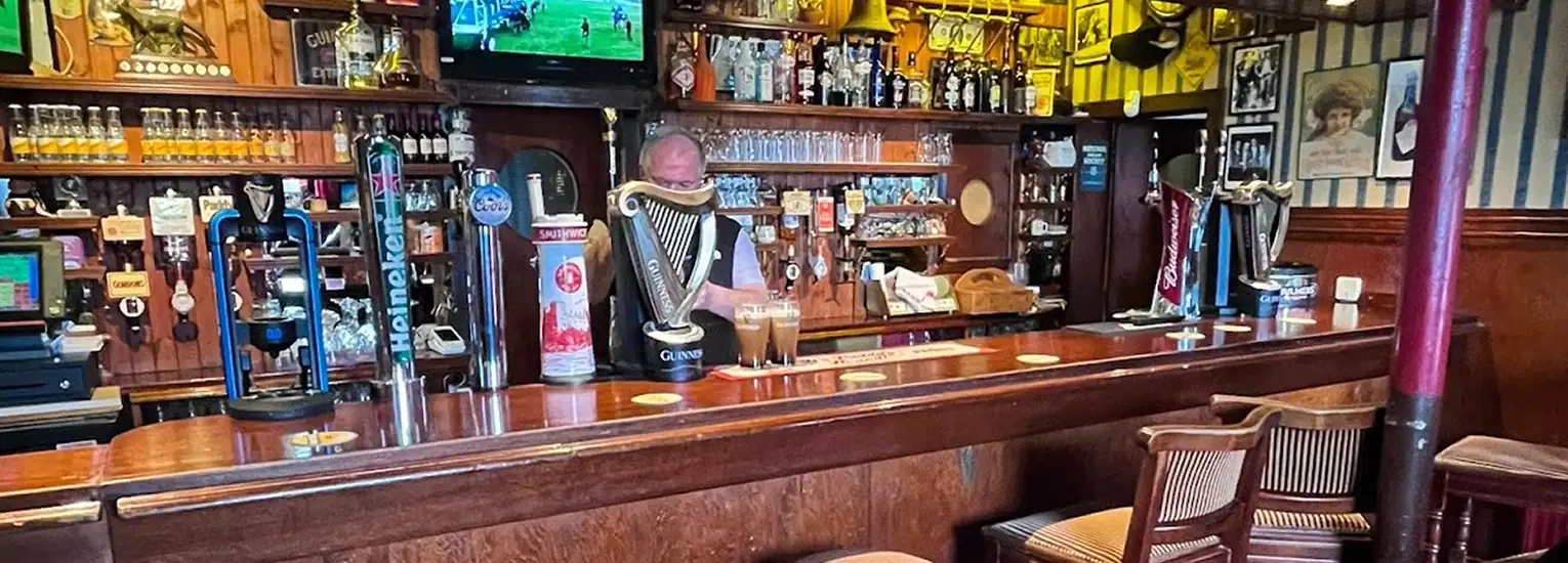 A barman stands behind a wooden bar in a cosy pub, with beer taps, glasses, and bottles visible. A telly above shows a sports match, and empty chairs line the bar counter.