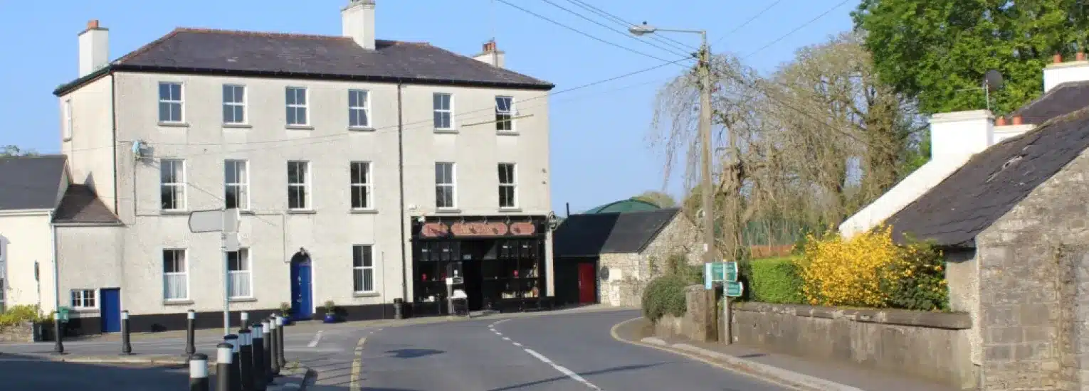 A quiet village street curves past a large, pale three-storey building with a shop on the ground floor, surrounded by stone buildings, greenery, and a clear blue sky.