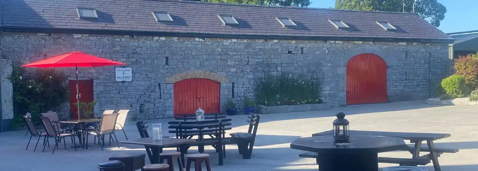 Outdoor seating area with tables, chairs, and a red umbrella in front of a stone building with red doors and several small windows on the roof, under a clear blue sky.