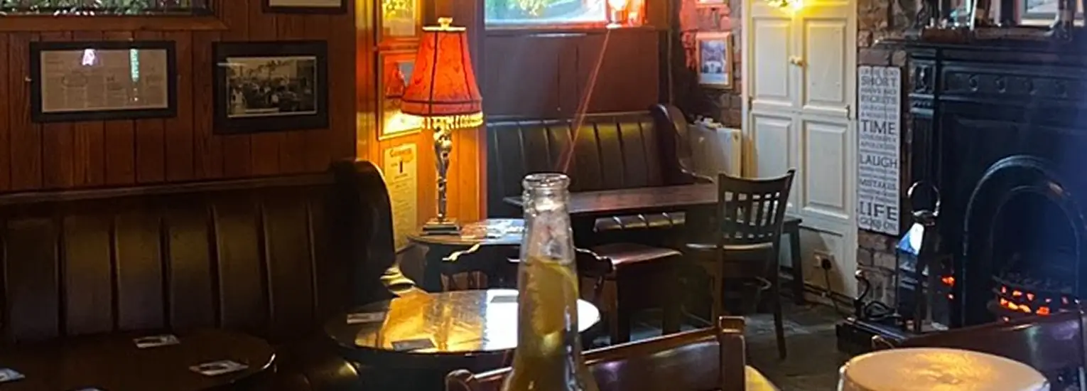 A cosy pub interior with wooden panelling, dark booths, a glowing fireplace, framed pictures on the walls, a lamp on a table, and drinks on the tables, including a bottle and a pint of beer in the foreground.