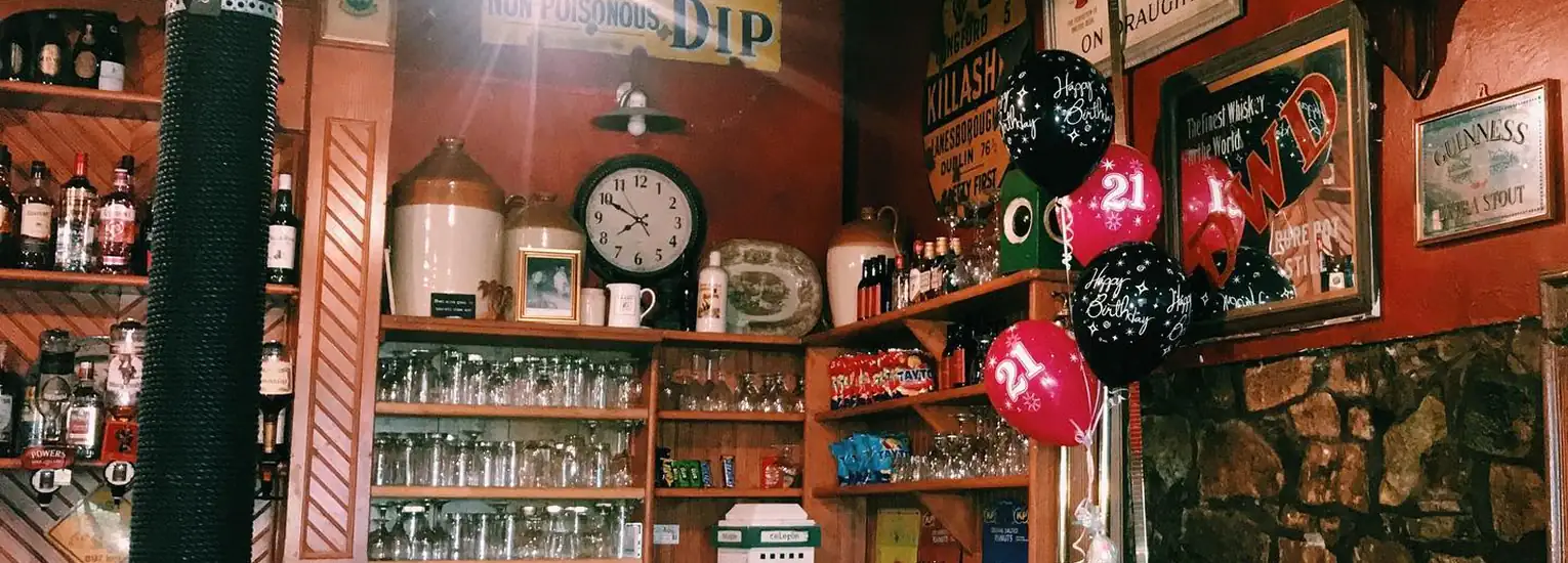 A cosy bar with shelves of glasses, bottles, and snacks, a wall clock, and black and pink "21" birthday balloons. Warm lighting highlights vintage decor and framed posters on the walls.