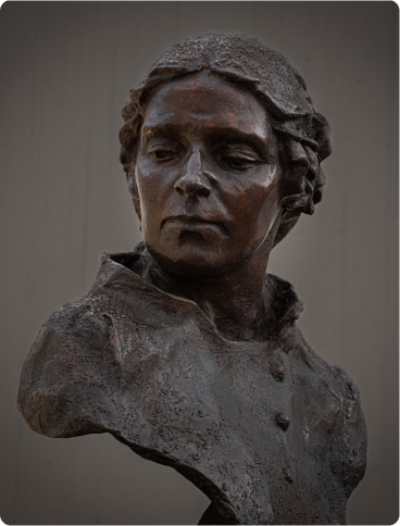 Bronze bust of a woman with plaited hair, wearing a high-collared garment, looking slightly down and to her left against a neutral background.