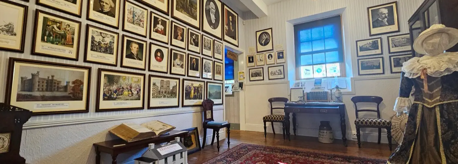 A museum room with framed portraits and historical images on the walls, antique chairs, a vintage desk with old items, a mannequin in period costume, and a window letting in natural light.