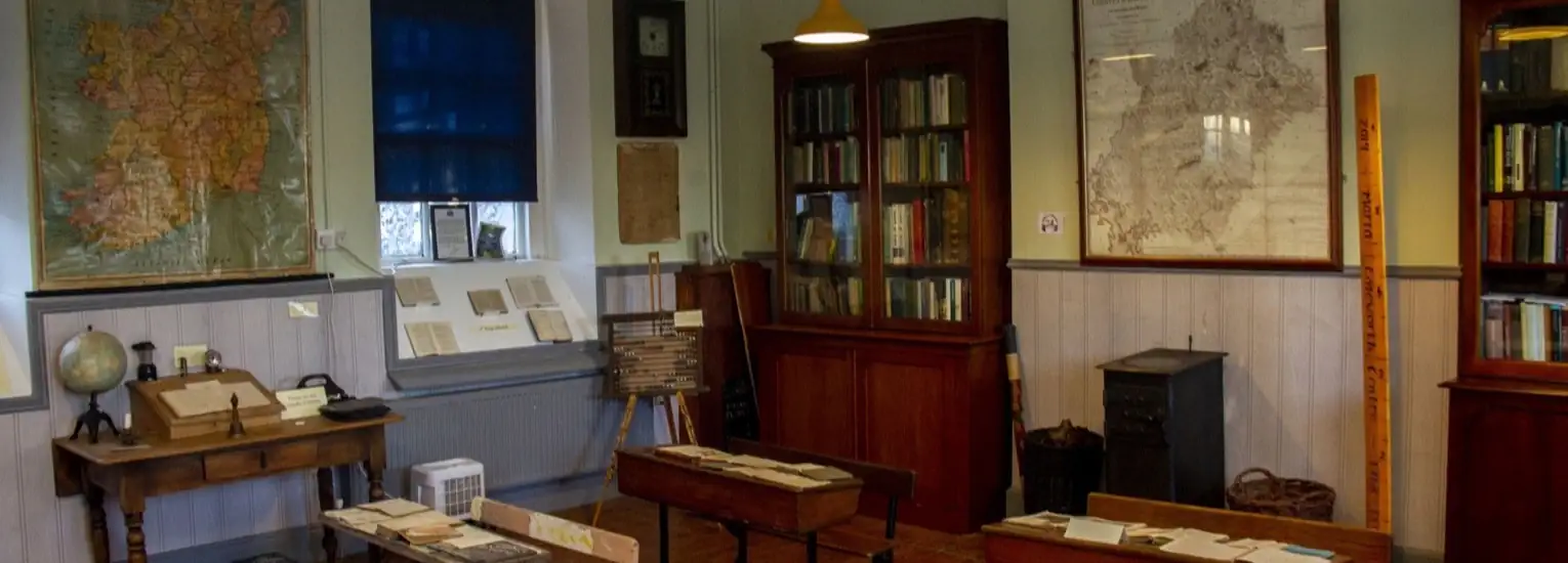A vintage classroom with wooden desks, a globe, and bookshelves. Large maps hang on the walls, and books and papers are arranged neatly. The room has warm lighting and an old-fashioned atmosphere.
