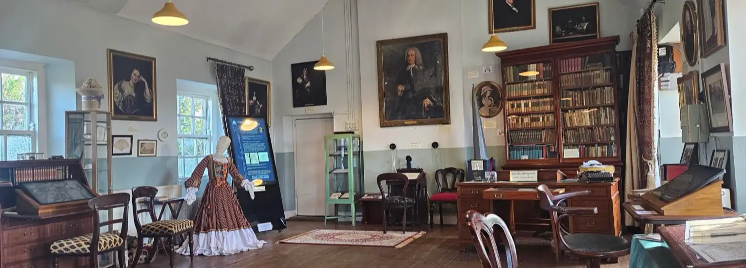 An historic room with antique furniture, bookshelves filled with books, framed portraits on the walls, a mannequin in a period dress, and various artefacts displayed throughout.