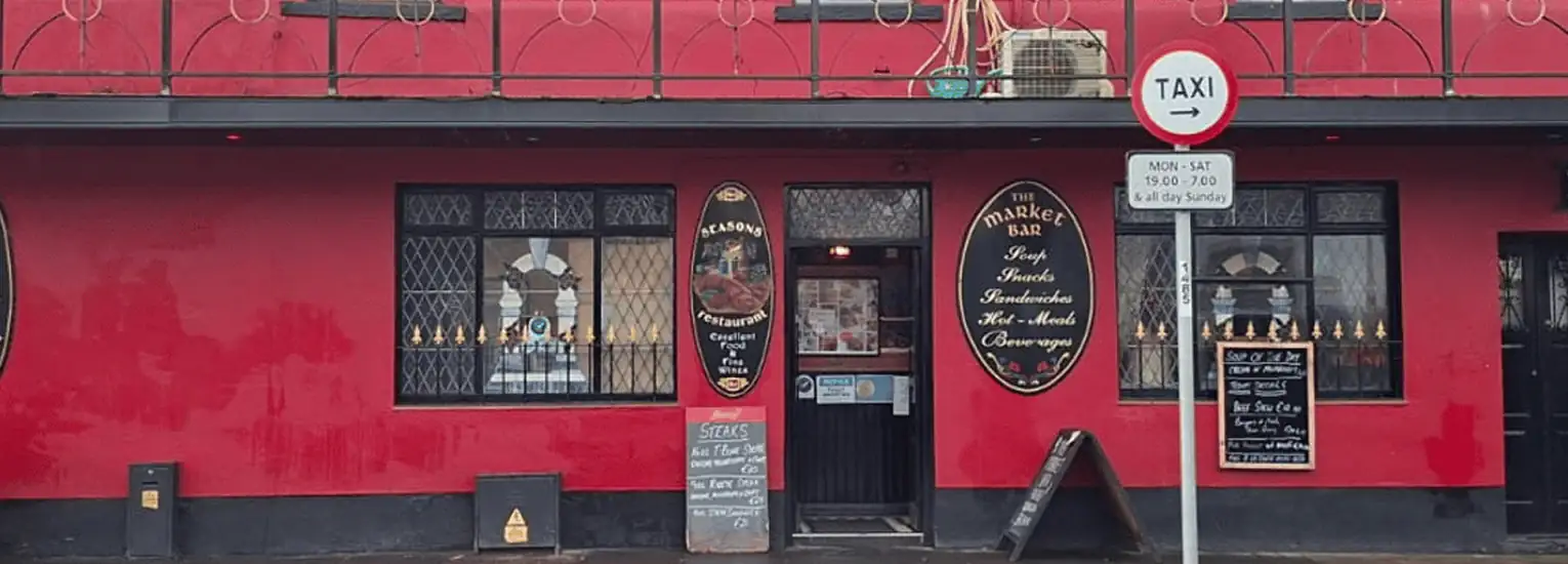 A bright red pub exterior with black trim, barred windows, and oval signs reading "The Drake Bar." A "TAXI" road sign stands outside, and chalkboards advertise specials near the entrance door.