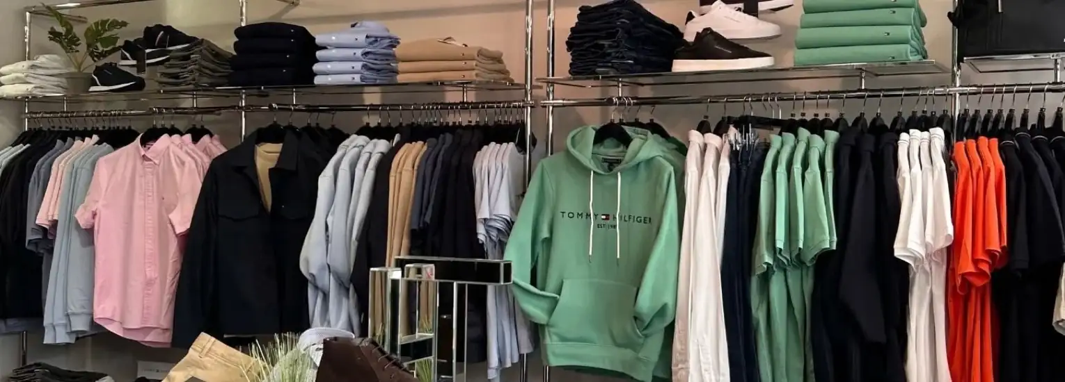 Clothing shop interior with neatly arranged shirts, hoodies, and trousers on racks and shelves. Display includes folded jeans, stacked shoes, and a green hoodie in the centre. The setting is organised and modern.