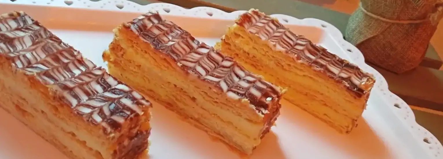 Three rectangular slices of layered pastry with a white and chocolate zigzag icing pattern on top, arranged neatly on a white decorative tray.