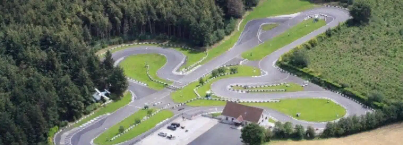 Aerial view of a go-kart racing track with winding asphalt lanes, bordered by green grass and trees. A small building with a car park is visible near the track’s entrance.