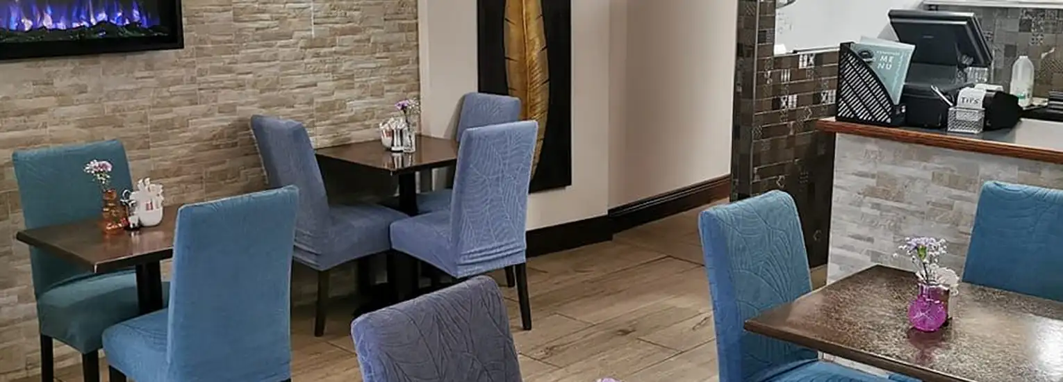 A cosy restaurant interior with blue upholstered chairs, dark wooden tables, small vases with flowers, a digital fireplace on a stone wall, and a front counter with a cash register.