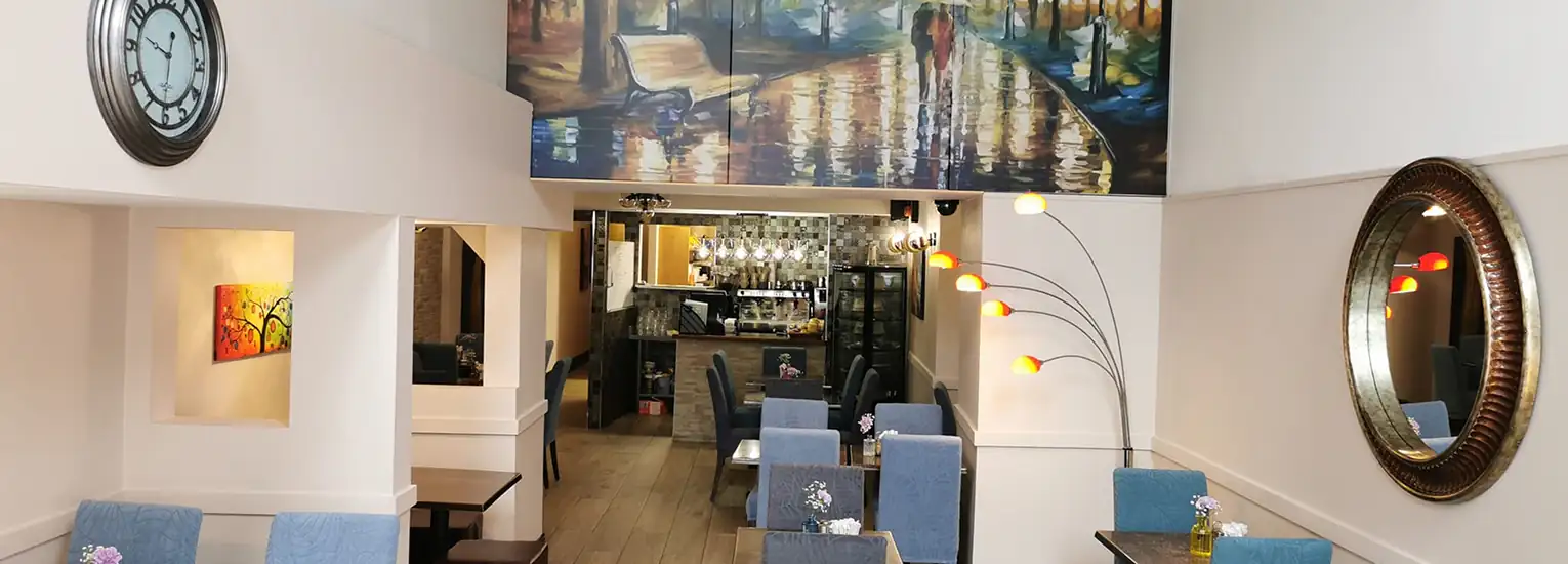 A cosy restaurant interior with blue upholstered chairs, small tables, a large wall clock, a round mirror, colourful wall art, and a corner floor lamp with multiple yellow and orange lights. The back shows a bar area.