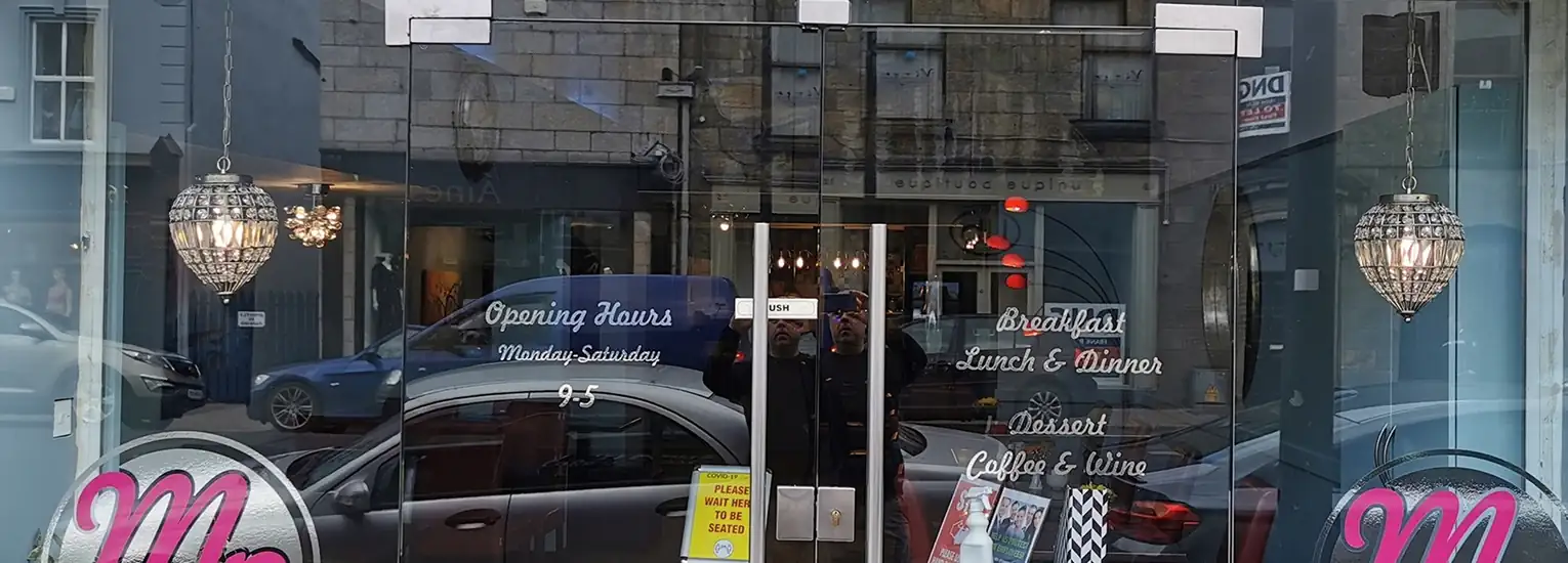 A glass-fronted café with hanging chandeliers, visible tables and chairs inside, and signs listing opening times and menu items like breakfast, lunch, dinner, dessert, coffee, and wine. Cars are parked outside.