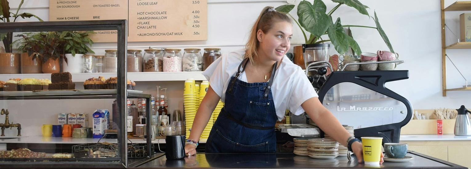 A smiling barista in a café stands behind the counter, reaching for a yellow coffee cup. Cakes and pastries are displayed in a glass case, and a menu board hangs on the wall in the background.