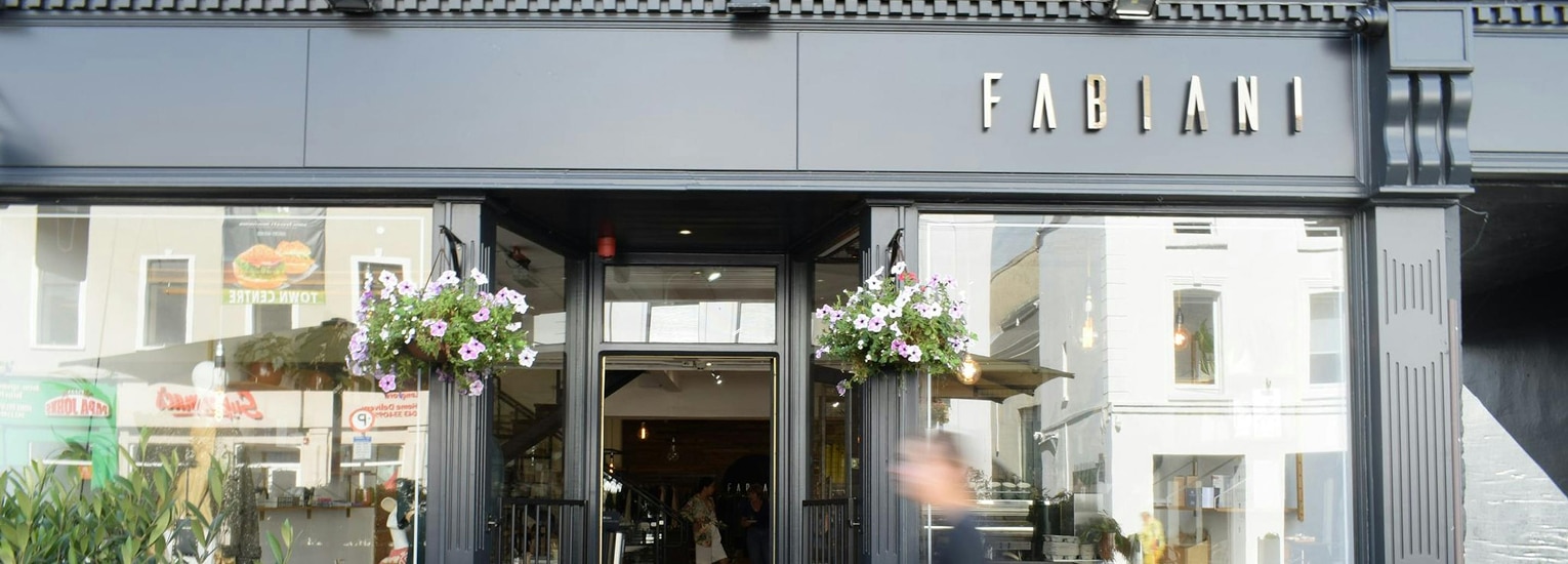 Street view of the entrance to Fabiani, a modern shopfront with large glass windows, black exterior, hanging flower baskets, and the Fabiani sign above the door. A person is walking past the entrance.