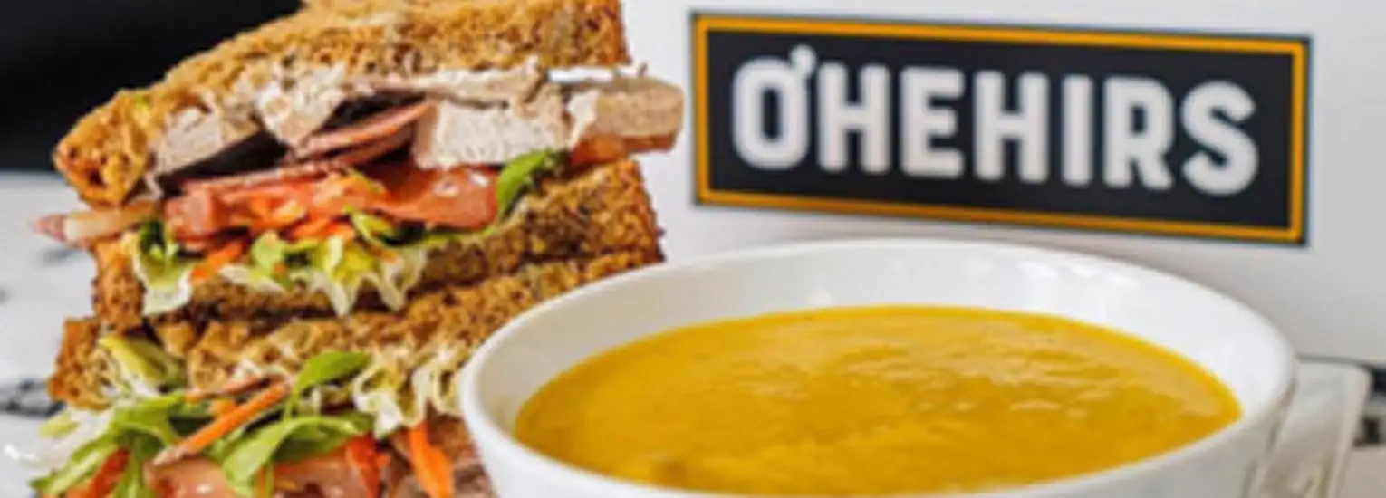 A bowl of creamy orange soup sits next to a stacked sandwich filled with turkey, lettuce, tomato, and other fresh vegetables. A sign in the background reads "O’Hehirs.