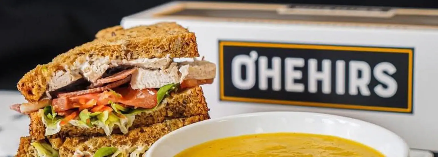 A sandwich with lettuce, tomato, and sliced meat on wholegrain bread sits next to a bowl of yellow soup, with an O’Hehirs-branded box in the background.
