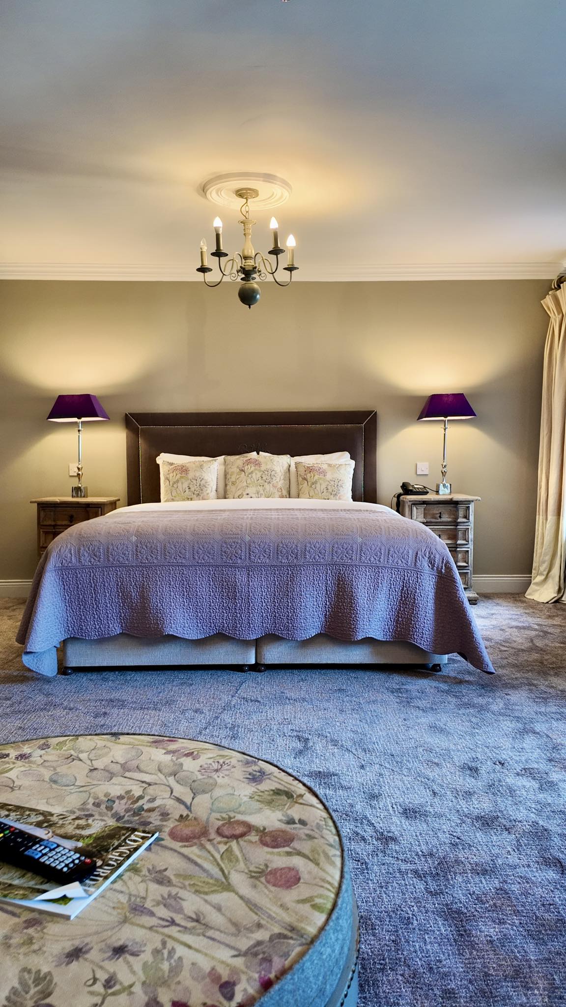 A cosy bedroom with a double bed, lavender bedspread, floral pillows, two bedside tables with purple lamps, a chandelier overhead, and a round, floral-patterned pouffe in the foreground.