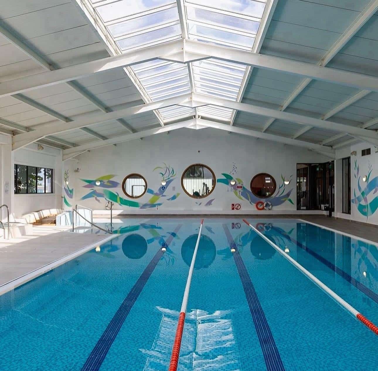 Indoor swimming pool with two swim lanes, blue tiled floor, and skylight ceiling. Wall features colourful fish murals and round windows. Sunlight shines through the glass roof, illuminating the bright, clean space.