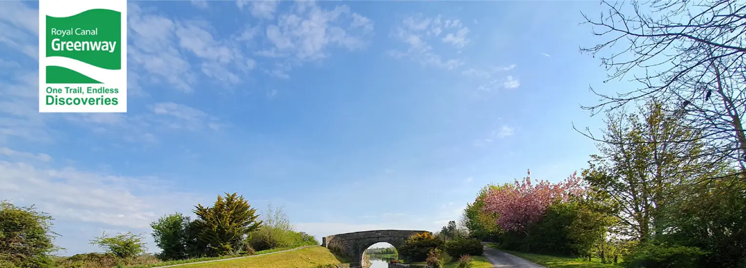 A scenic canal path bordered by trees and grass leads to a stone bridge under a blue sky. In the top left corner, a Royal Canal Greenway logo reads, "One Trail, Endless Discoveries.