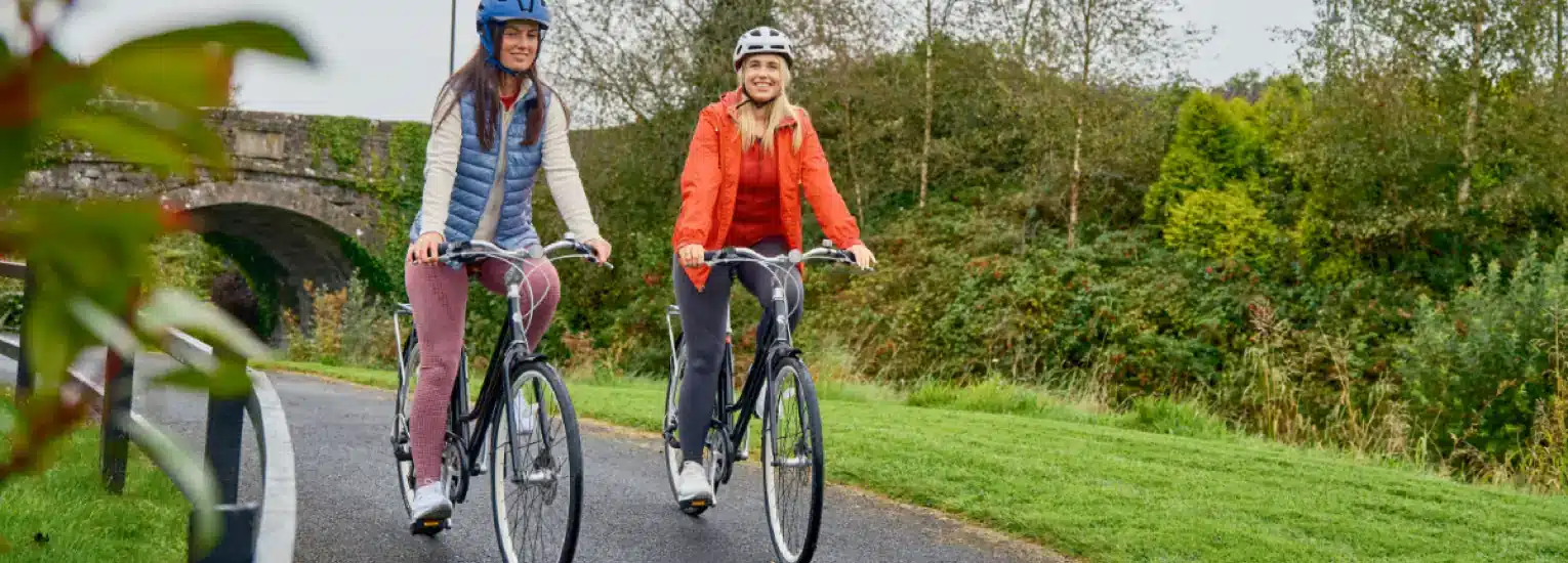 Two women wearing helmets and jackets ride bicycles on a paved path through a green park, with trees and a stone bridge in the background. Both women are smiling and enjoying the outdoor activity.