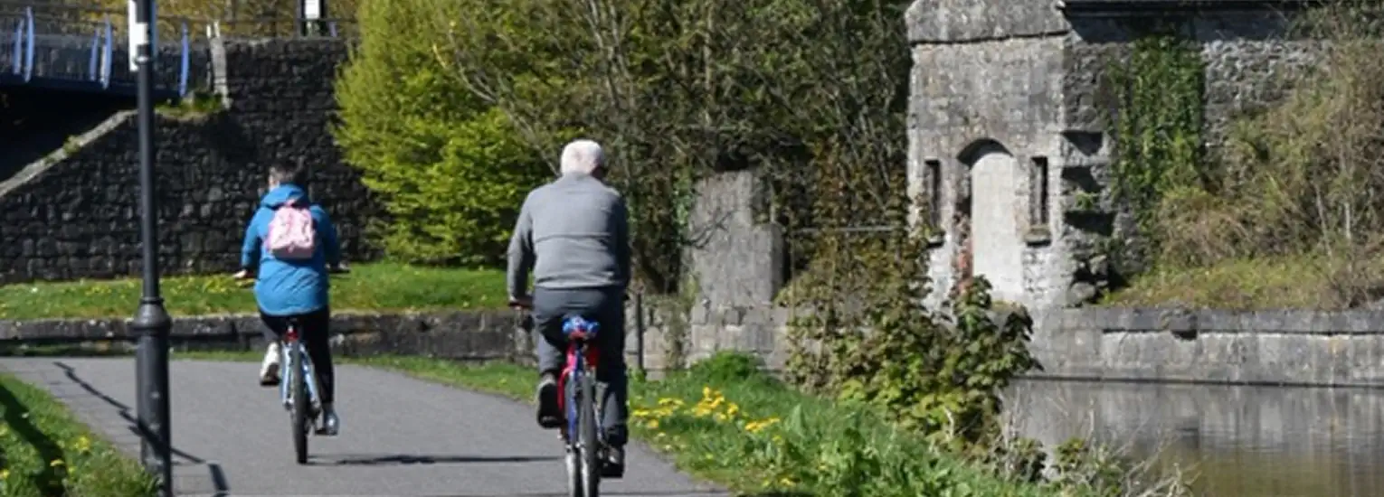 Two people ride bicycles along a paved path beside a canal with stone walls, greenery, and an old stone structure on the right. The scene is outdoors on a sunny day.