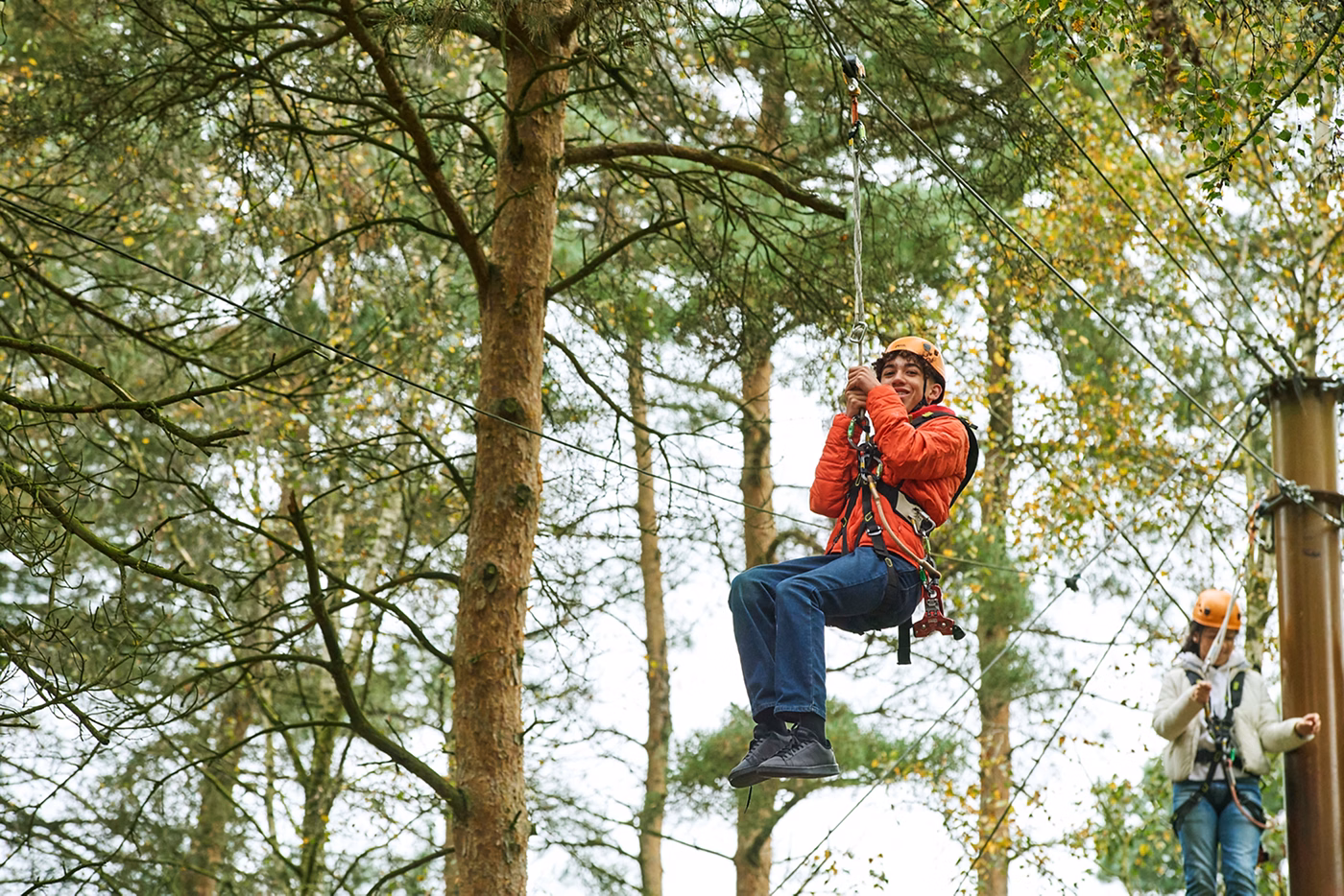A person wearing a helmet and harness is zip lining through a forest. Another person stands in the background on a wooden platform among tall trees. Both appear to be enjoying an outdoor adventure activity.