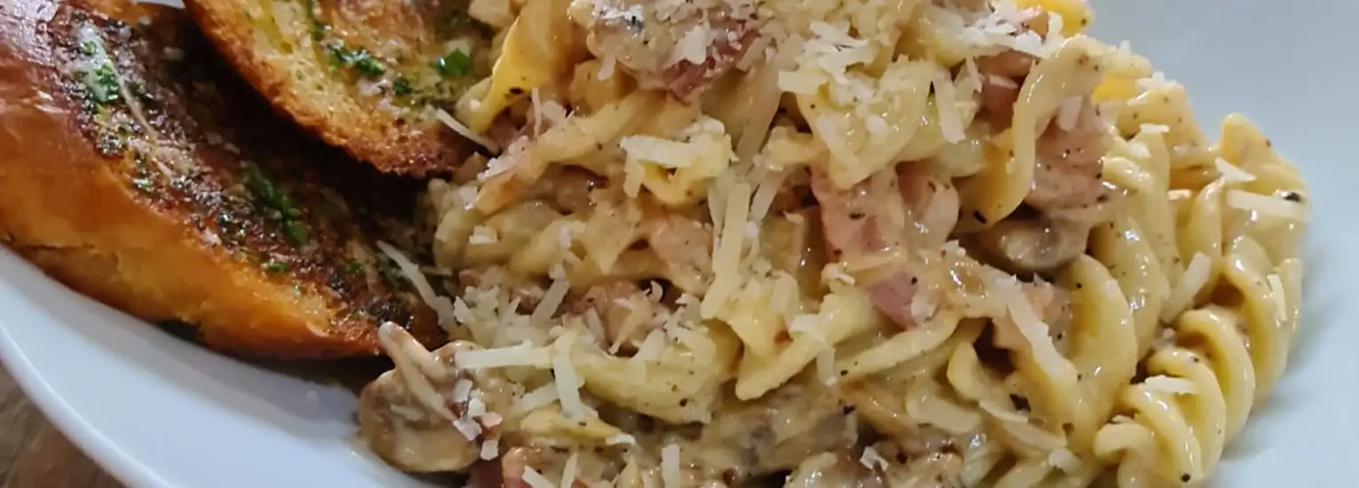 A close-up of creamy pasta with mushrooms and ham, topped with grated cheese, served with two slices of golden garlic bread on the side.