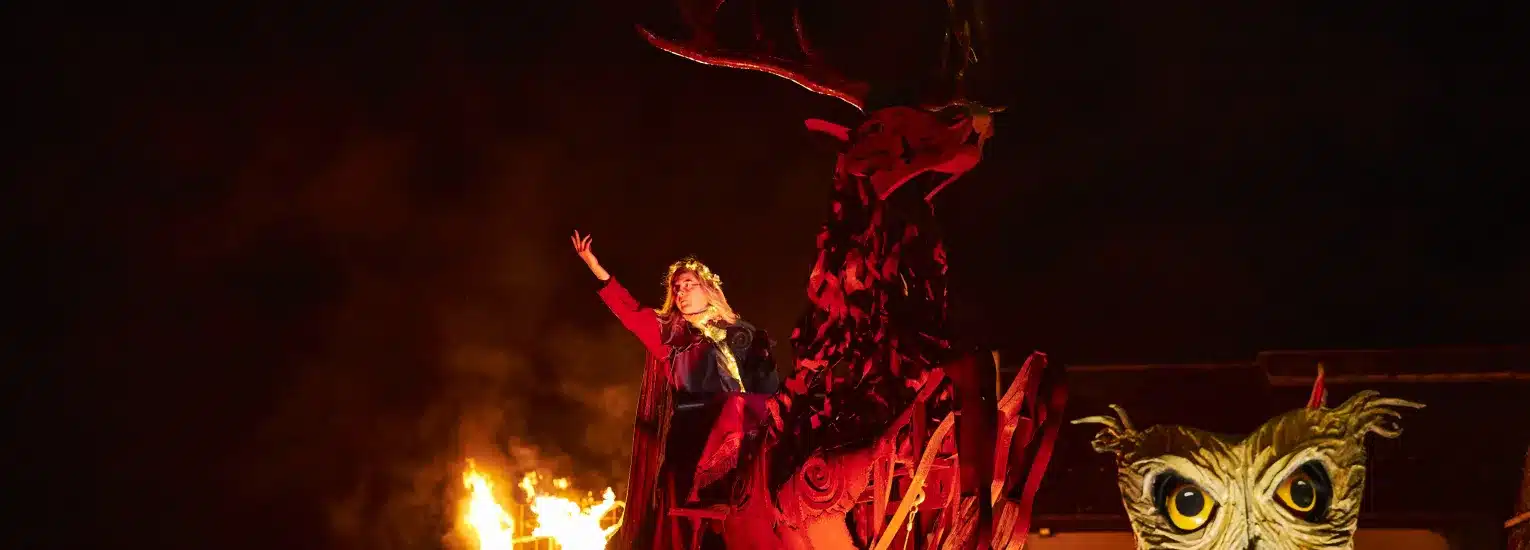 A person in a crown and red robe stands atop a large stag sculpture, raising one arm, with flames burning in the background and an owl mask visible in the lower right-hand corner.