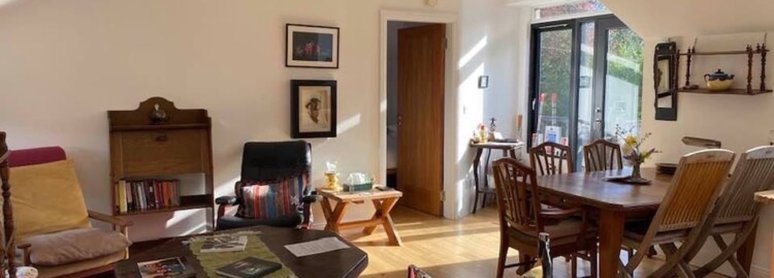 A sunlit living and dining room with wooden furniture, bookshelves, framed pictures on white walls, and a mix of chairs around a dining table. Light streams in from a glass door leading outside.