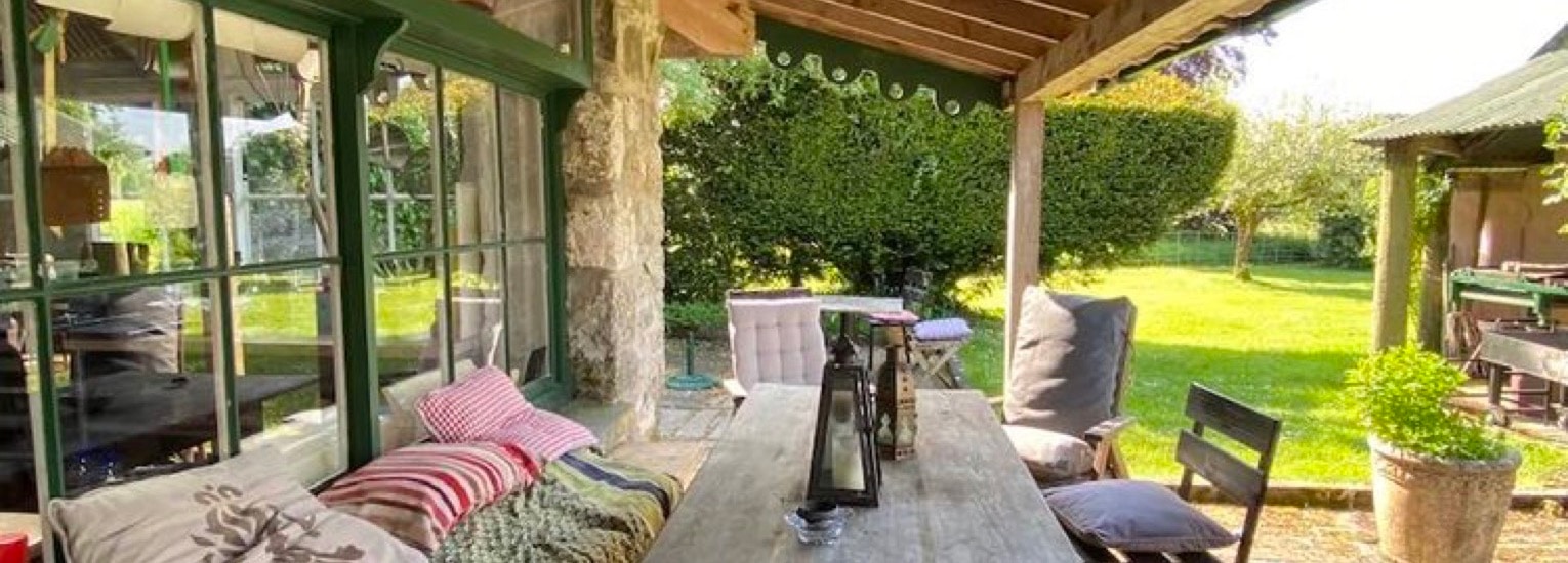 A cosy covered patio with cushioned seating, a wooden table, and chairs, overlooking a green garden. Sunlight brightens the space, and decorative cushions add a colourful, inviting touch.