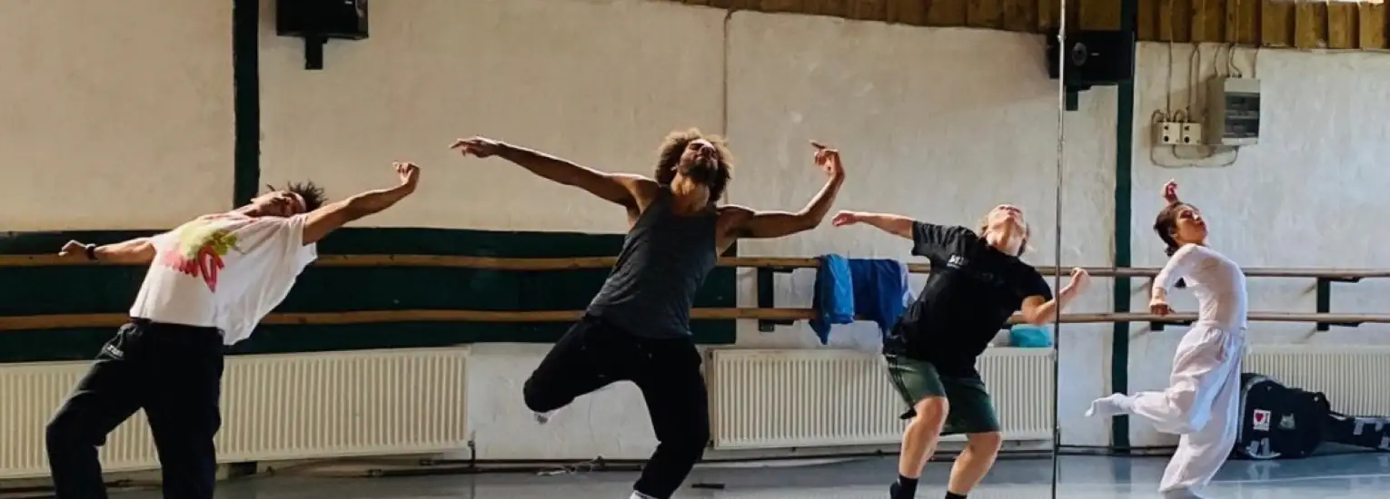 Four dancers, each in unique expressive poses, rehearse in a studio with mirrored walls, wooden barres, and jackets hanging in the background. Sunlight streams in from above, illuminating the energetic scene.