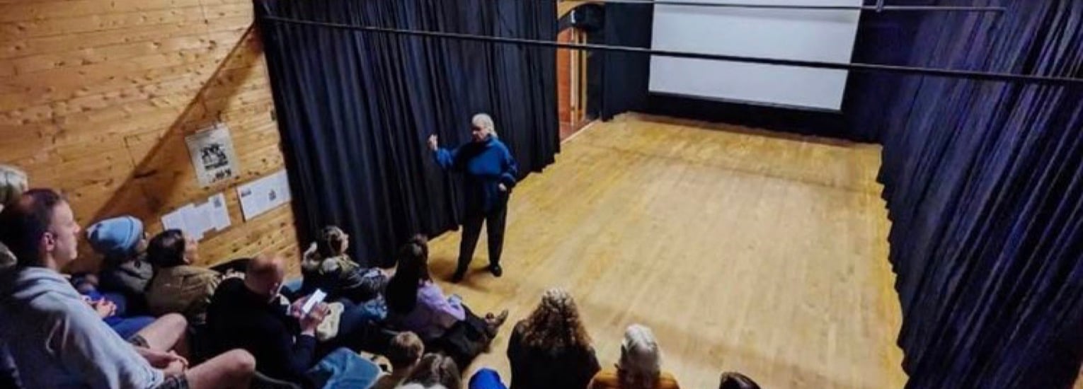 A person stands on a wooden stage in front of seated audience members, speaking or presenting. The room has black curtains, a screen at the back, and wooden walls.