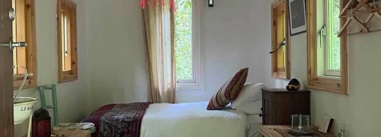 A cosy bedroom with a single bed, white bedding, and patterned blanket. There are wooden-framed windows, a wooden bedside table, shelves, and natural light streaming in, creating a warm and inviting atmosphere.