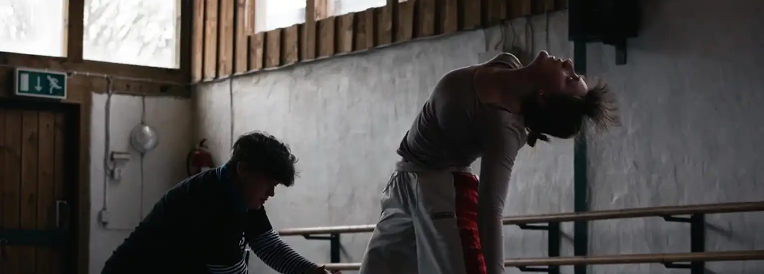 Two people in a dimly lit room; one person is sitting or kneeling and appears to be assisting or observing, whilst the other bends backwards dramatically during what seems to be a dance or movement practice.