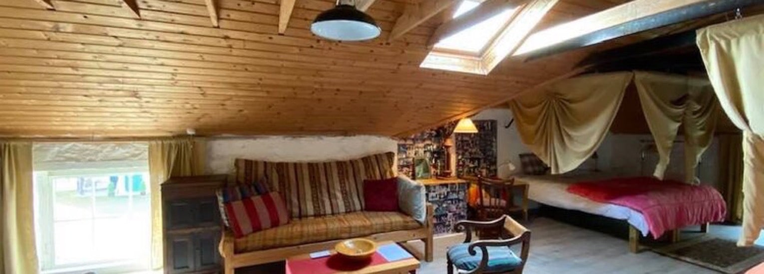 A cosy attic room with a sloped wooden ceiling, skylight, sofa with striped cushions, coffee table, armchair, and bed with a pink blanket; curtains partially divide the sleeping area. Warm natural light fills the space.