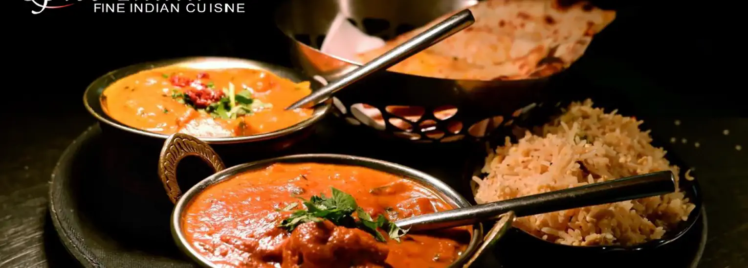 Two bowls of Indian curry with metal spoons, a serving of rice, and naan bread are arranged on a black plate. The food is garnished with herbs and spices, creating an inviting, colourful meal.