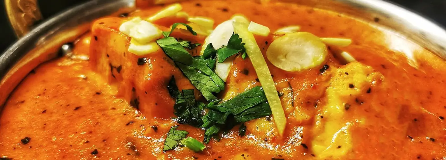 Close-up of a dish with paneer cubes in a creamy orange curry sauce, garnished with chopped coriander, sliced green chillies, and ginger strips, served in a metal bowl.