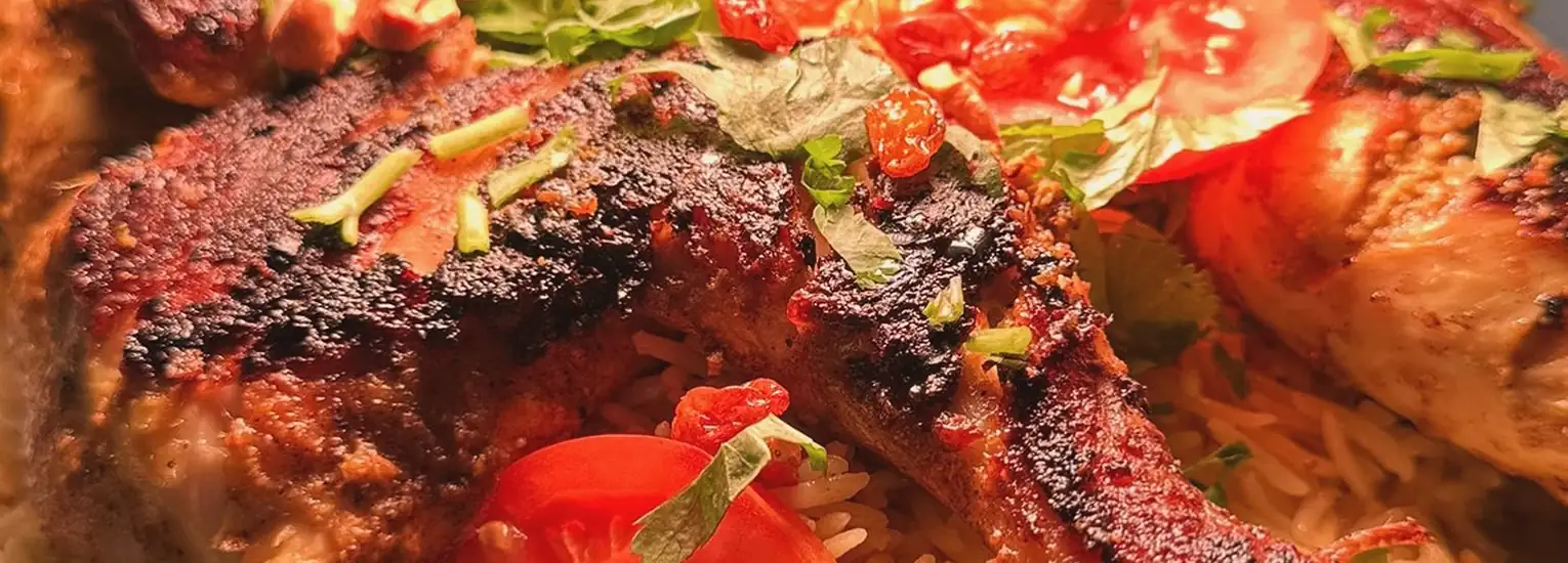 Close-up of grilled chicken legs served on a bed of rice, garnished with sliced tomatoes and fresh herbs. The chicken appears well-seasoned with a charred, crispy exterior.
